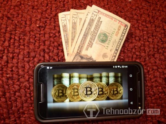 Монеты Биткоина на экране смартфона, лежащего на 20-долларовых купюрах
