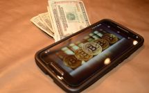 Смартфон с Биткоинами на дисплее лежит на 20-долларовых банкнотах